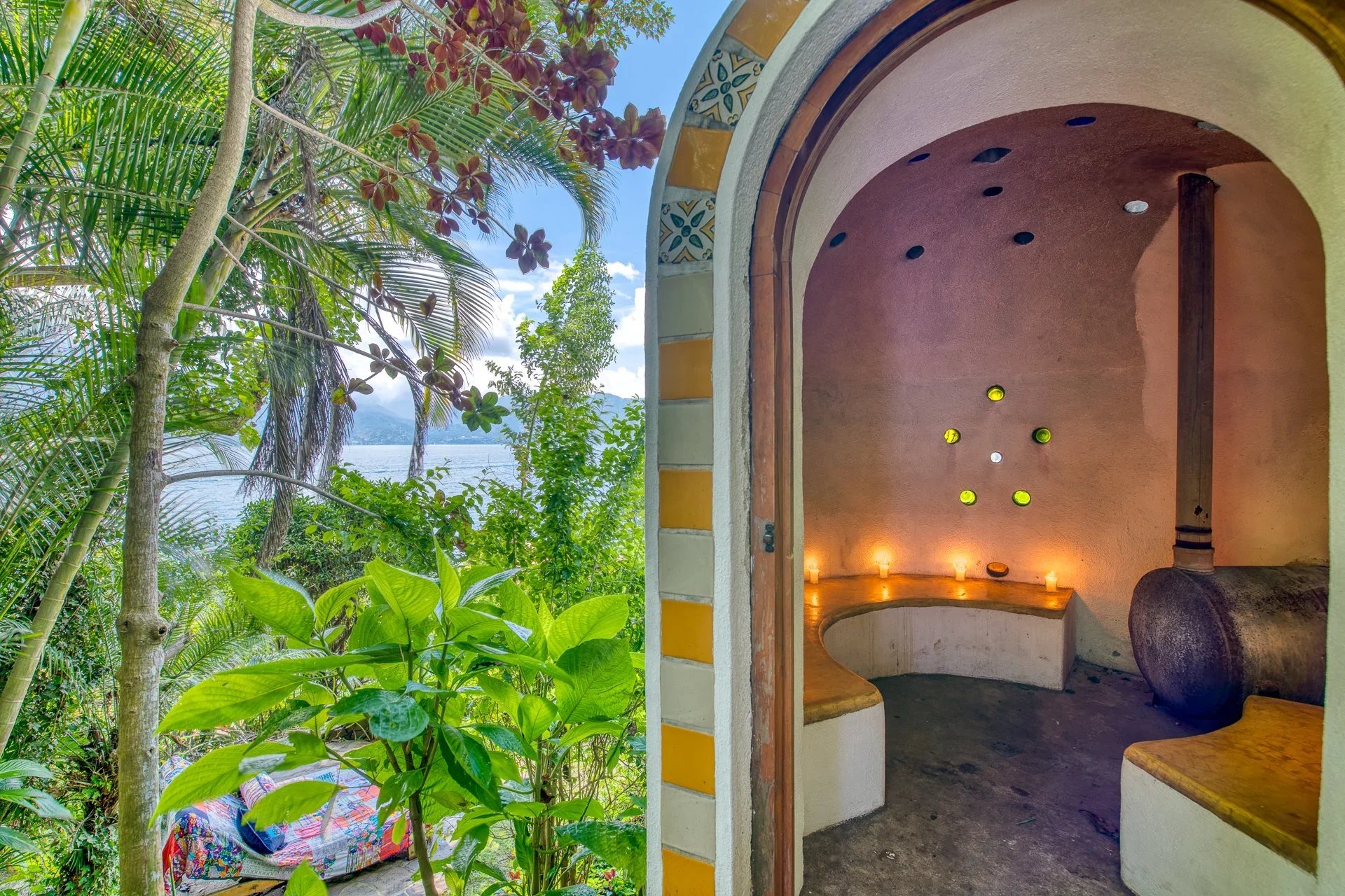 View of a lush tropical garden with trees and plants on the left, and a small interior room with rounded walls, candles, and decorative wall lights on the right.