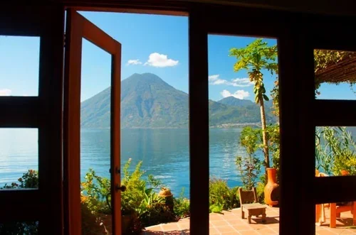 View through open door and window showing a lake with mountains in the background, trees, and outdoor patio furniture.