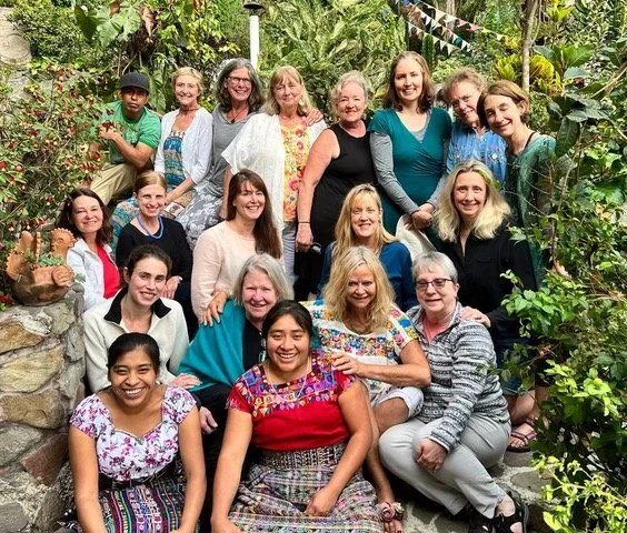 Group of 16 women and one man posing together outdoors in a lush garden with green plants and trees, some standing and others sitting on rocks.
