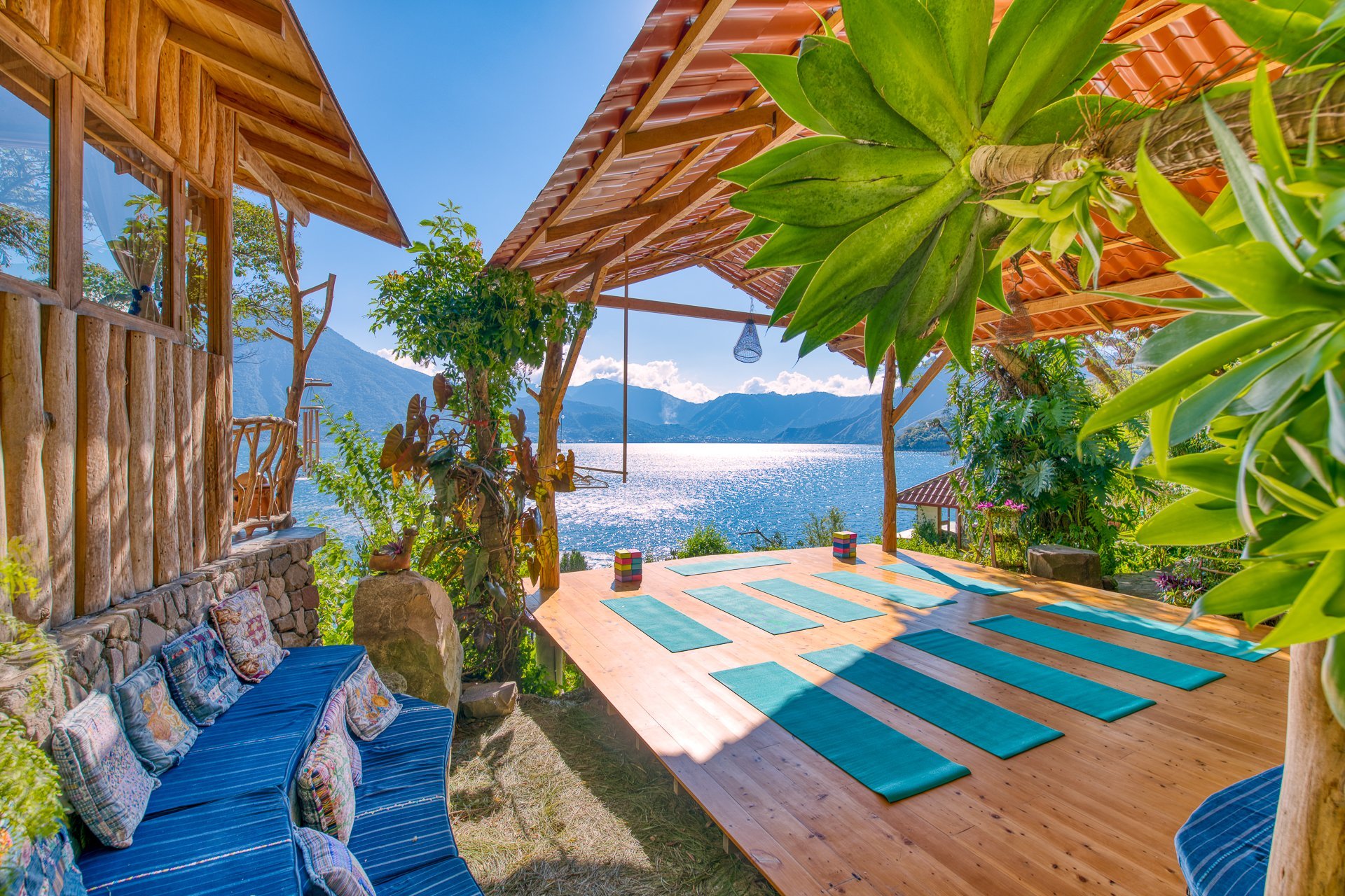 Outdoor wooden deck with yoga mats, surrounded by green plants and trees, overlooking a lake and mountains under a blue sky.