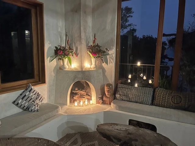 Cozy living room corner with built-in bench seating, decorative pillows, and a fireplace with candles and floral arrangements on top. Large windows show trees and outdoor lights at night.