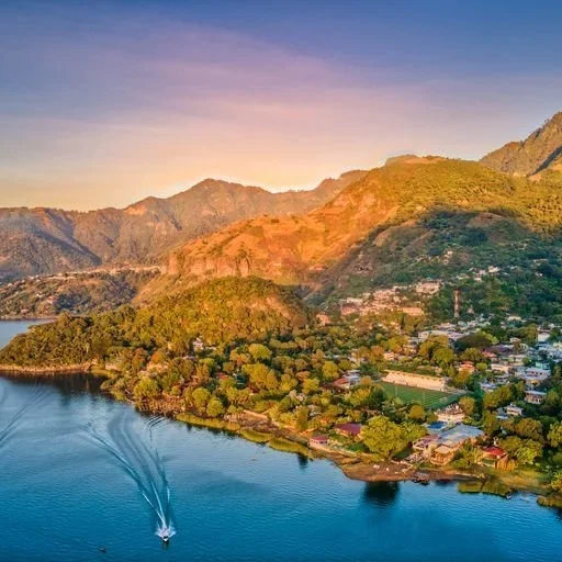 Aerial view of a river surrounded by lush green trees and a small town with homes and buildings, with mountains in the background during sunset.