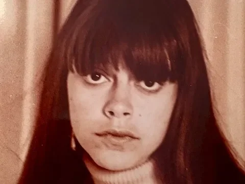 Young woman with long brown hair and bangs, wearing a turtleneck sweater, looking slightly to the side.