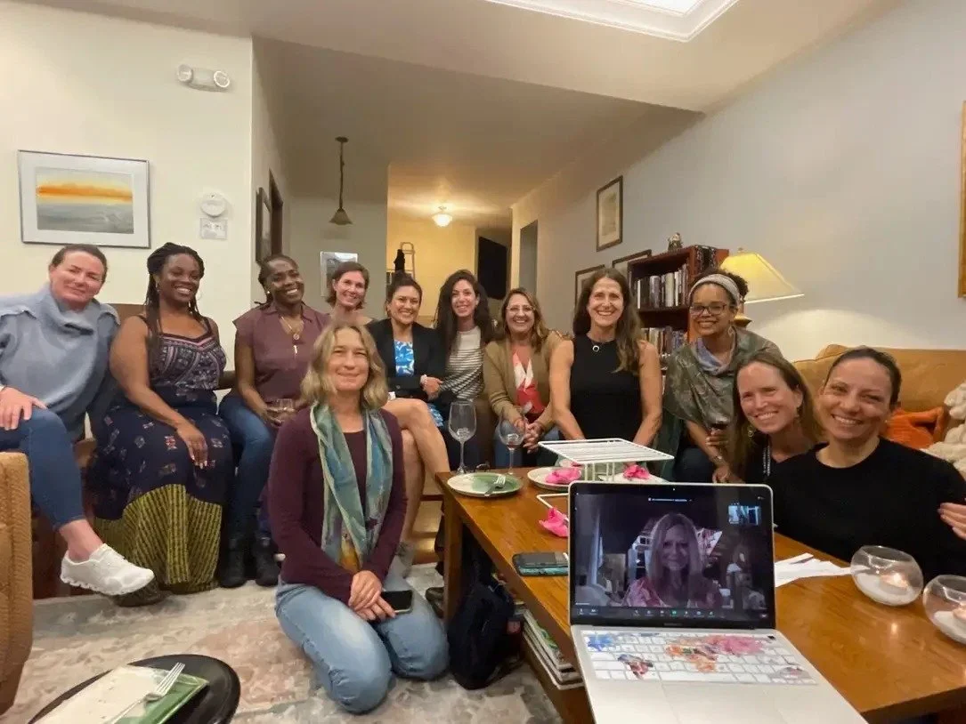 Group of women gathered in a living room, sitting and standing around a coffee table, some holding wine glasses, smiling for a photo with a woman on a video call on a laptop in the foreground.