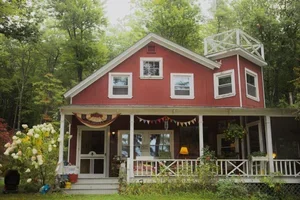 Red house with white trim, large front porch, surrounded by green trees and plants, with an upper balcony.