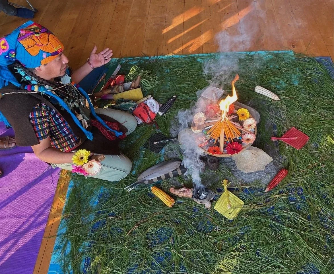 A person dressed in traditional indigenous attire sitting in front of a sacred fire, surrounded by offerings and natural items on a grassy mat.