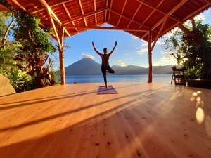 Person practicing yoga on a wooden platform with a mountain and lake in the background, under a roofed structure with trees on the sides.