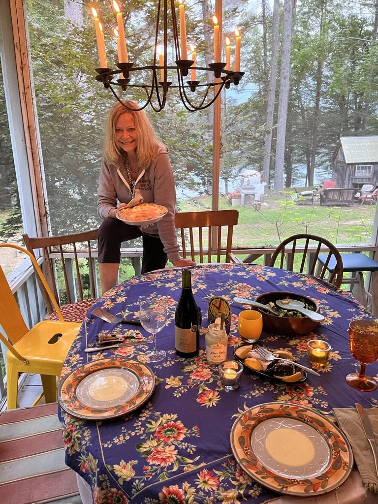 Woman standing by a round dining table with a floral tablecloth, holding a pizza. The table is set with plates, wine glasses, candles, and a bottle of wine. A chandelier hangs above, and a screened porch with a view of trees and a lake is in the background.