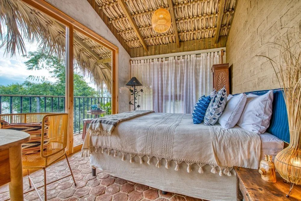 A cozy bedroom with a bed covered in a beige blanket and decorative pillows, large sliding glass door leading to a balcony with a rattan chair and a small table, tropical scenery outside, bamboo ceiling with a woven pendant light, wooden furniture, and earthy tones.