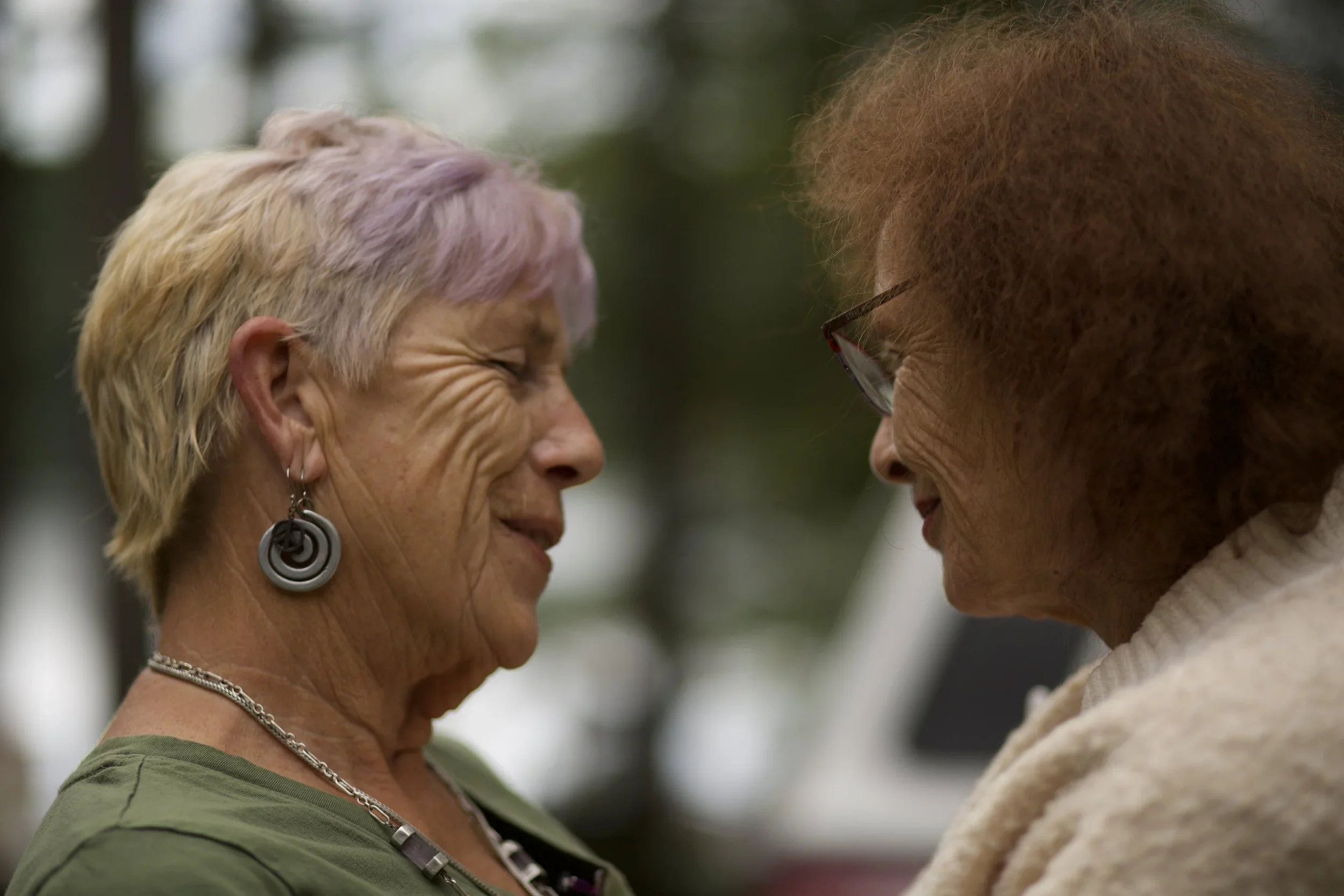 Two elderly women smiling and touching foreheads in an outdoor setting.