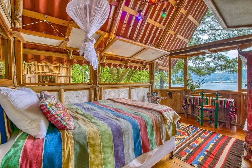 A cozy bedroom with a bamboo bed and rainbow-colored striped blanket, decorated with plaid pillows, in a rustic cabin overlooking a lake and surrounded by trees, with colorful string lights and a patterned rug.