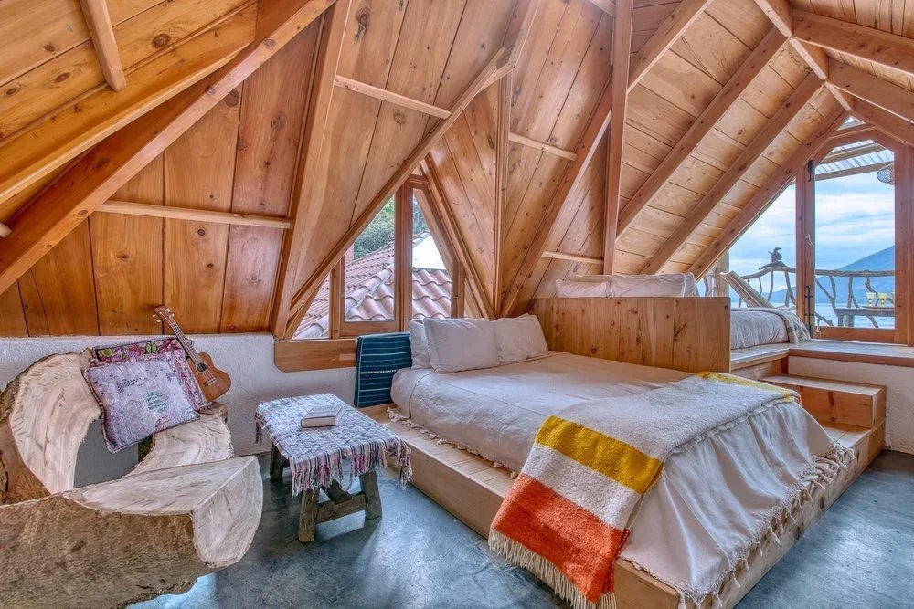 A cozy A-frame attic bedroom with wooden walls and ceiling, featuring a large bed with white linens, a colorful striped blanket, and a smaller bed near a window with a view of mountains and trees. There is a rustic wooden bench, a small side table with a book, and a guitar leaning against the wall.