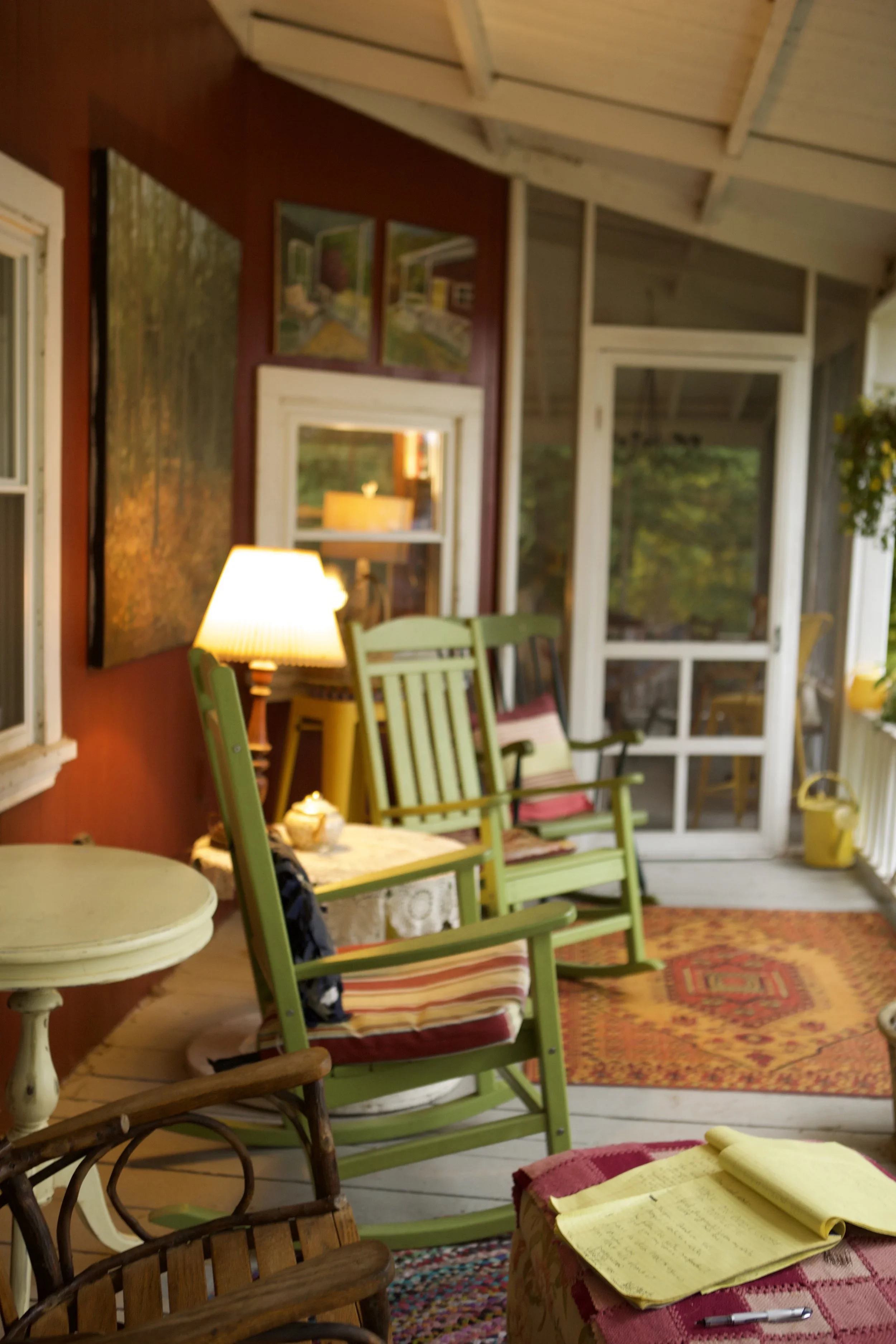A screened porch area with green rocking chairs, a small side table with a lamp, and colorful rugs. There are windows and artwork on the walls and a view of trees outside.