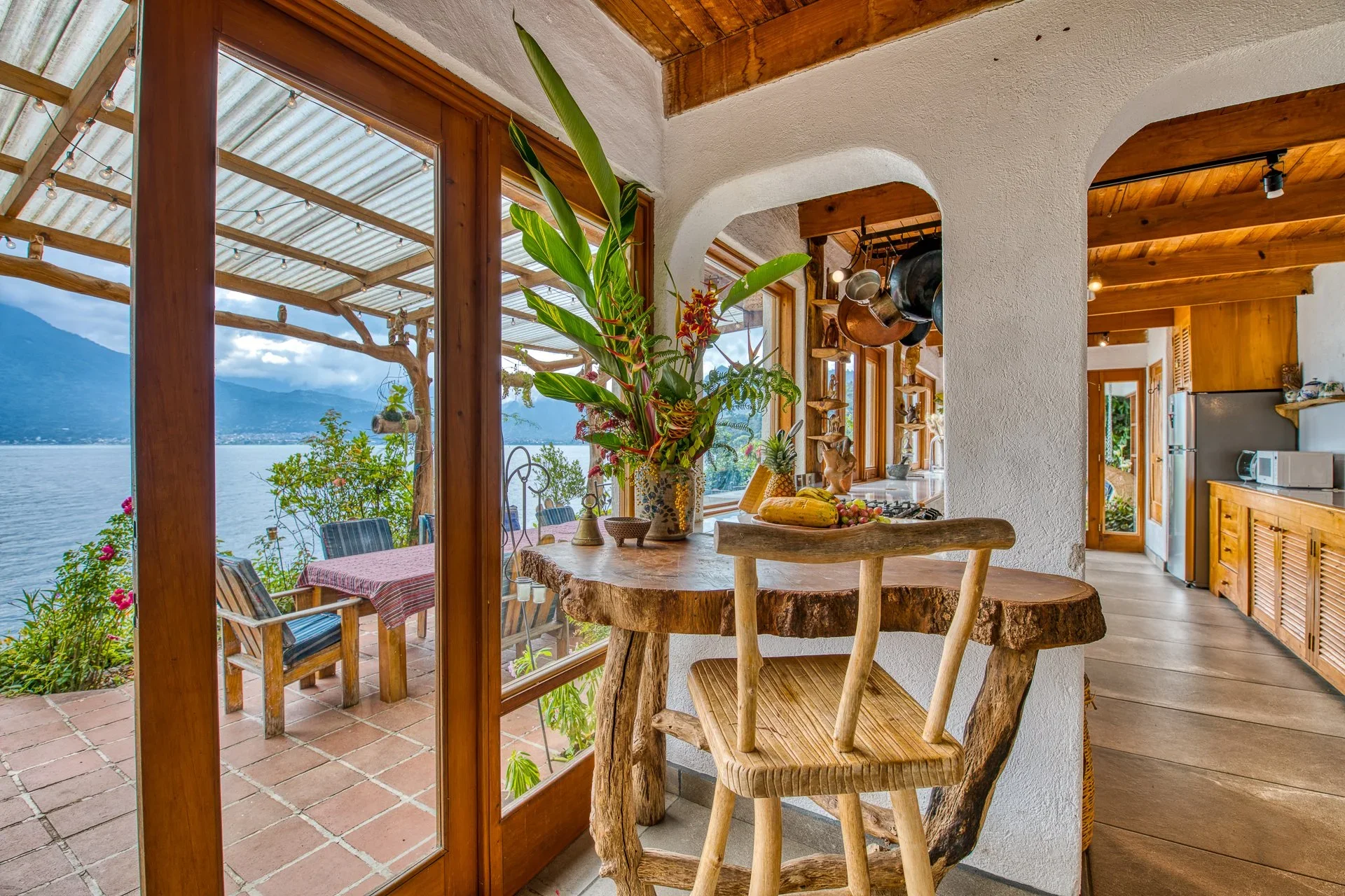 A cozy kitchen and dining area with a large window overlooking a lake and mountains, decorated with potted plants, wooden furniture, and hanging pots.