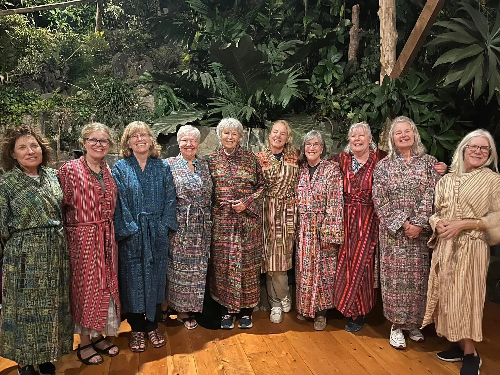 Group of women dressed in colorful striped robes standing together on wooden floor in front of lush tropical plants.