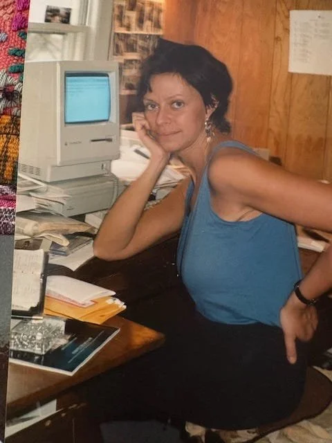 Woman with short dark hair in a blue sleeveless top sitting at cluttered desk with old computer and papers, in a wood-paneled office.