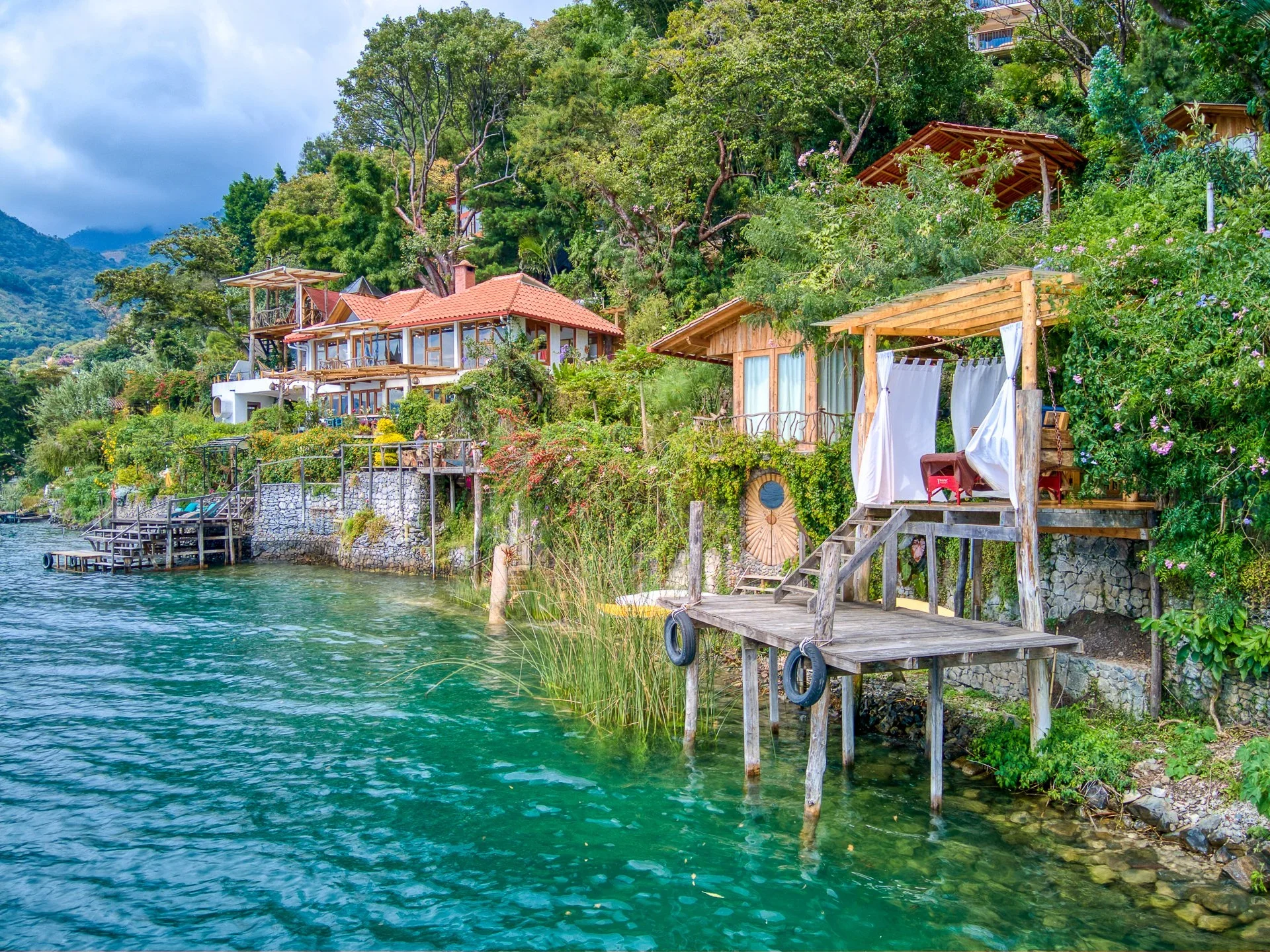 Colorful waterfront homes with wooden decks and lush greenery along a clear blue lake. Some homes have terraces overlooking the water, surrounded by trees and flowering bushes, with mountains in the background.