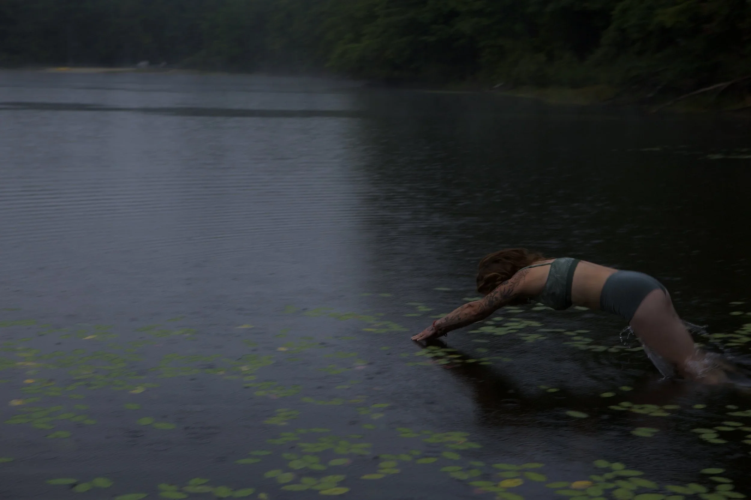 A woman with tattoos on her arms wearing a sports bra and shorts, leaning forward in a lake surrounded by trees, reaching into the water.