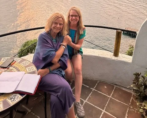 An older woman and a young girl sitting on a terrace overlooking a body of water, holding hands and smiling at the camera.