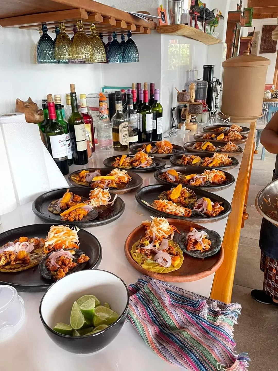 Several plates of food, possibly Mexican tacos, with shredded meat, onions, and lemon wedges, are arranged on a kitchen counter. Behind the counter are bottles of wine and liquor, and various kitchen items.