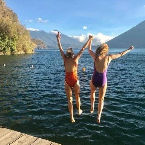 Two women jumping into the lake with mountains and blue sky in the background.