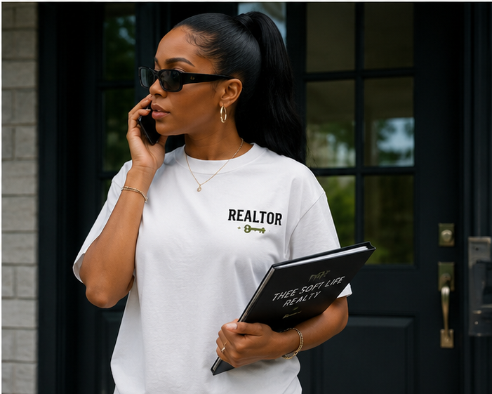 A woman wearing black sunglasses and a white T-shirt with 'REALTOR' written on it, standing outside near a house, talking on her phone and holding a notebook.