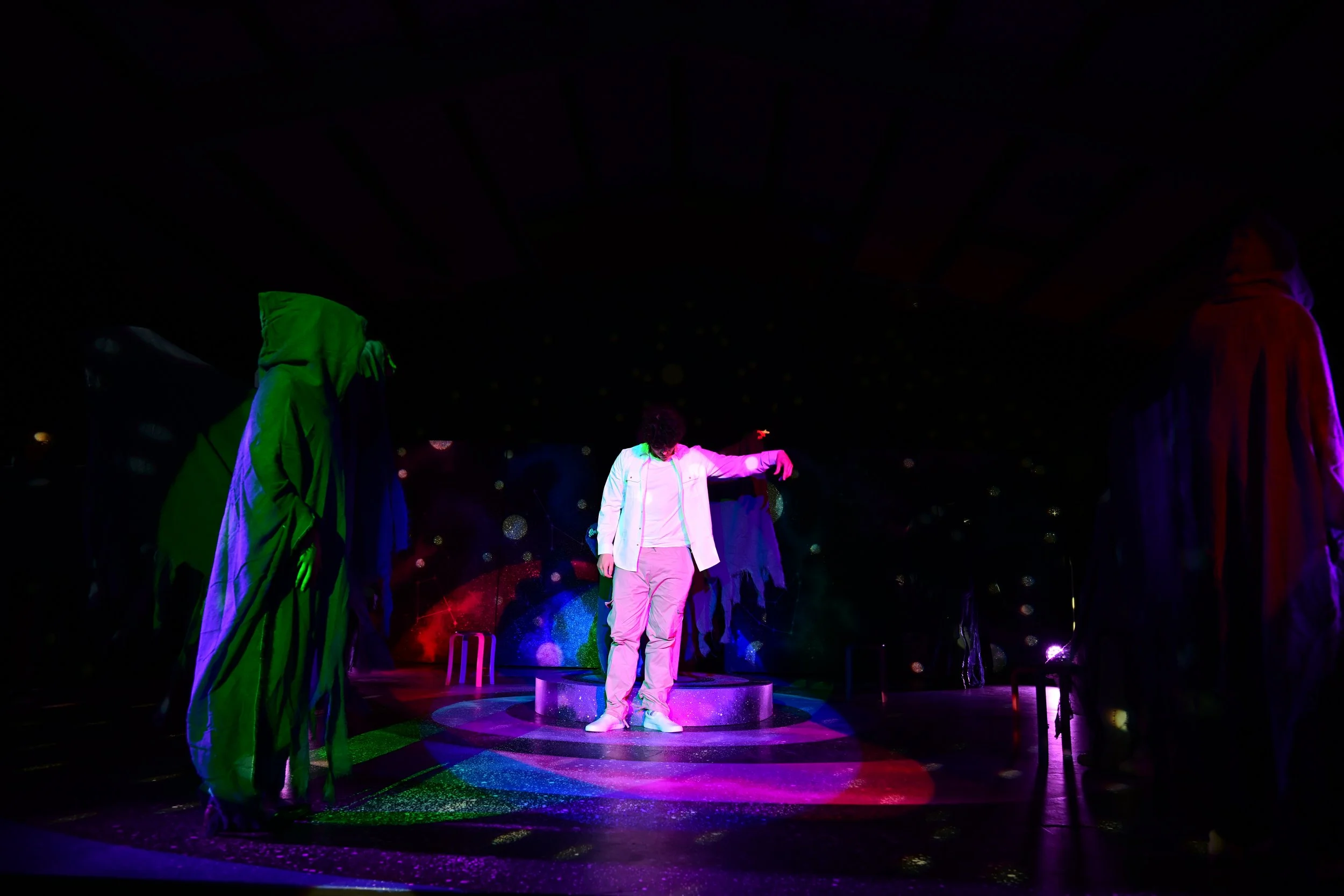 A person in white clothing on a small circular stage, surrounded by three figures in hooded cloaks with colorful lighting in a dark setting.