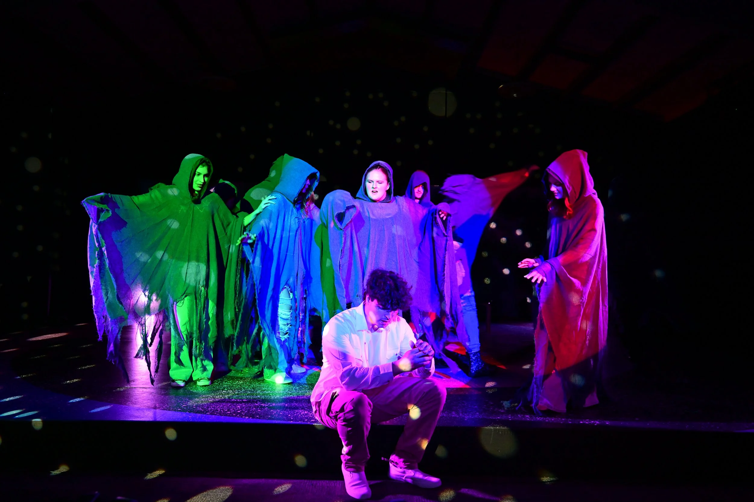 Group of people in colorful cloaks performing on stage with dark background and illuminated by stage lights.