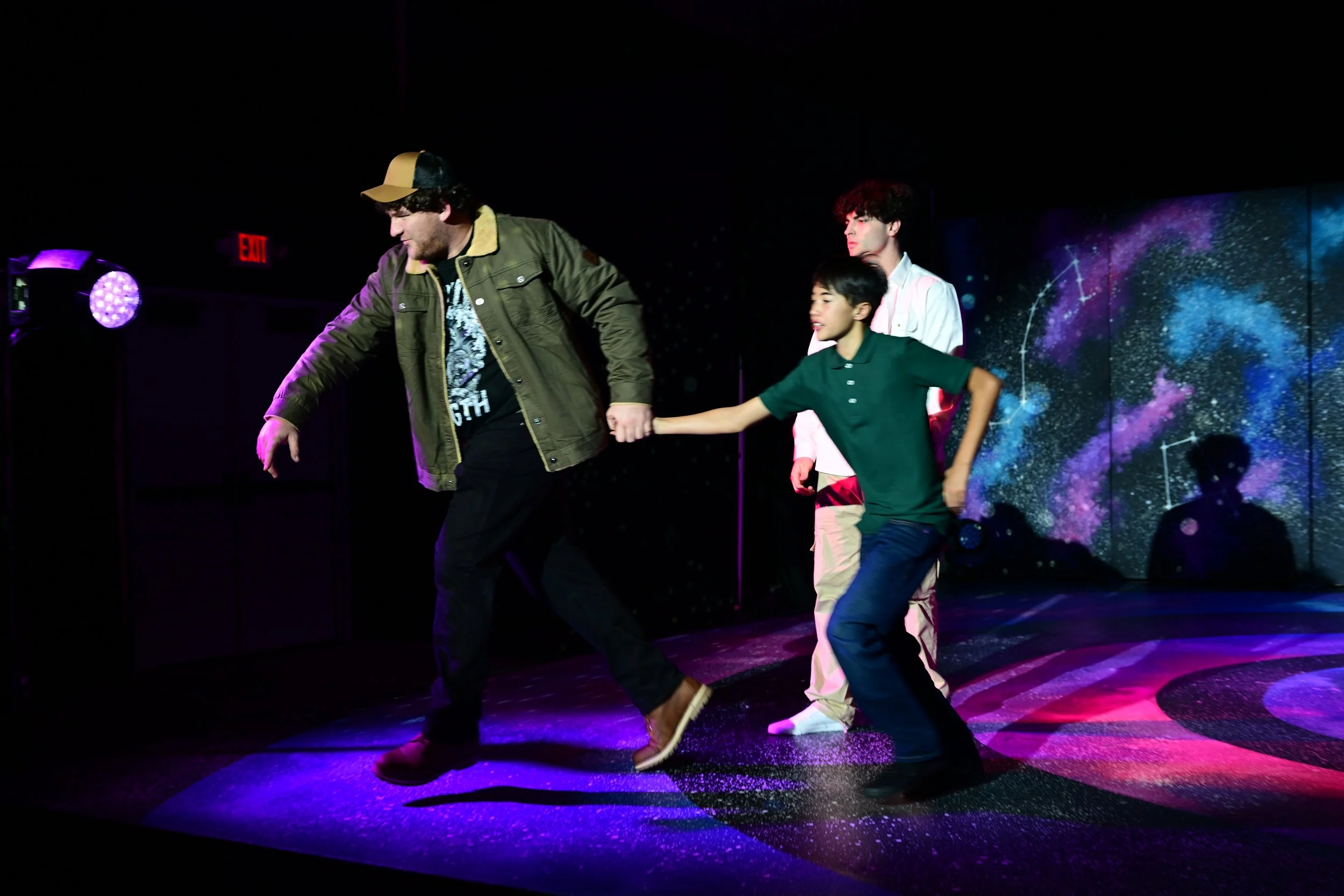 Three young men on stage, one holding the hand of another. The background features a colorful galaxy projection, and purple stage lighting is visible.