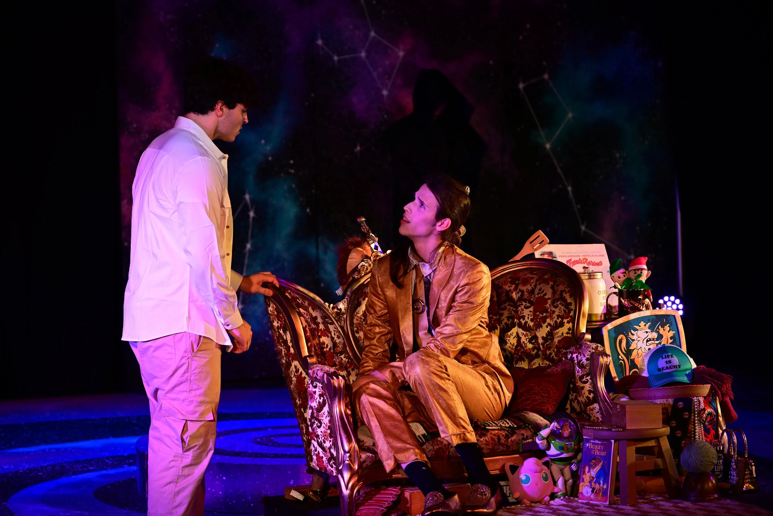A scene from a theatrical play showing a man in a white shirt and beige pants standing and talking to a woman in a golden suit sitting on an ornate vintage sofa. The background has a starry night and constellation design, and the scene is decorated w