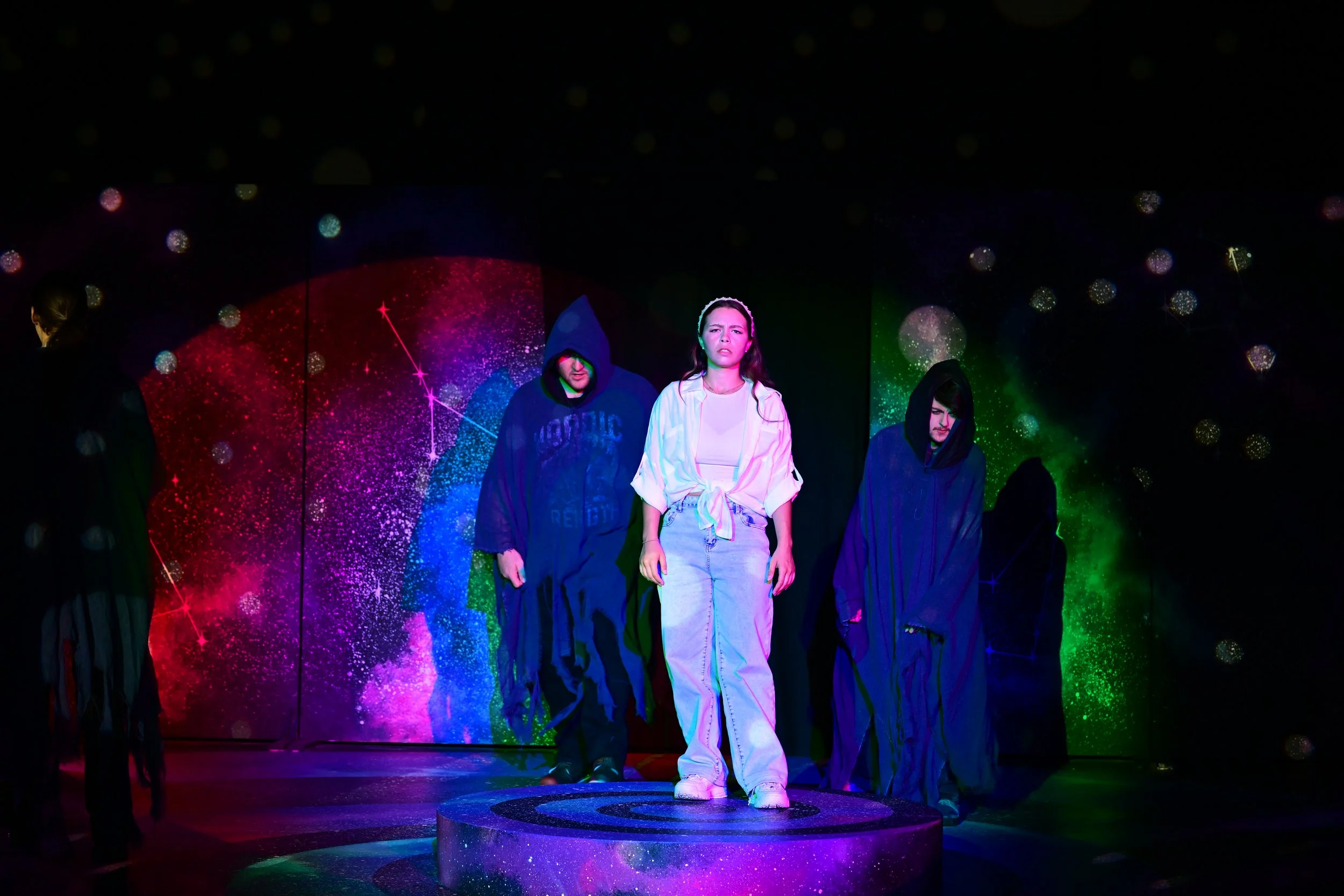 A young woman stands on a circular platform, surrounded by people dressed in dark hooded robes, with a colorful cosmic backdrop featuring constellations and nebulae behind her.