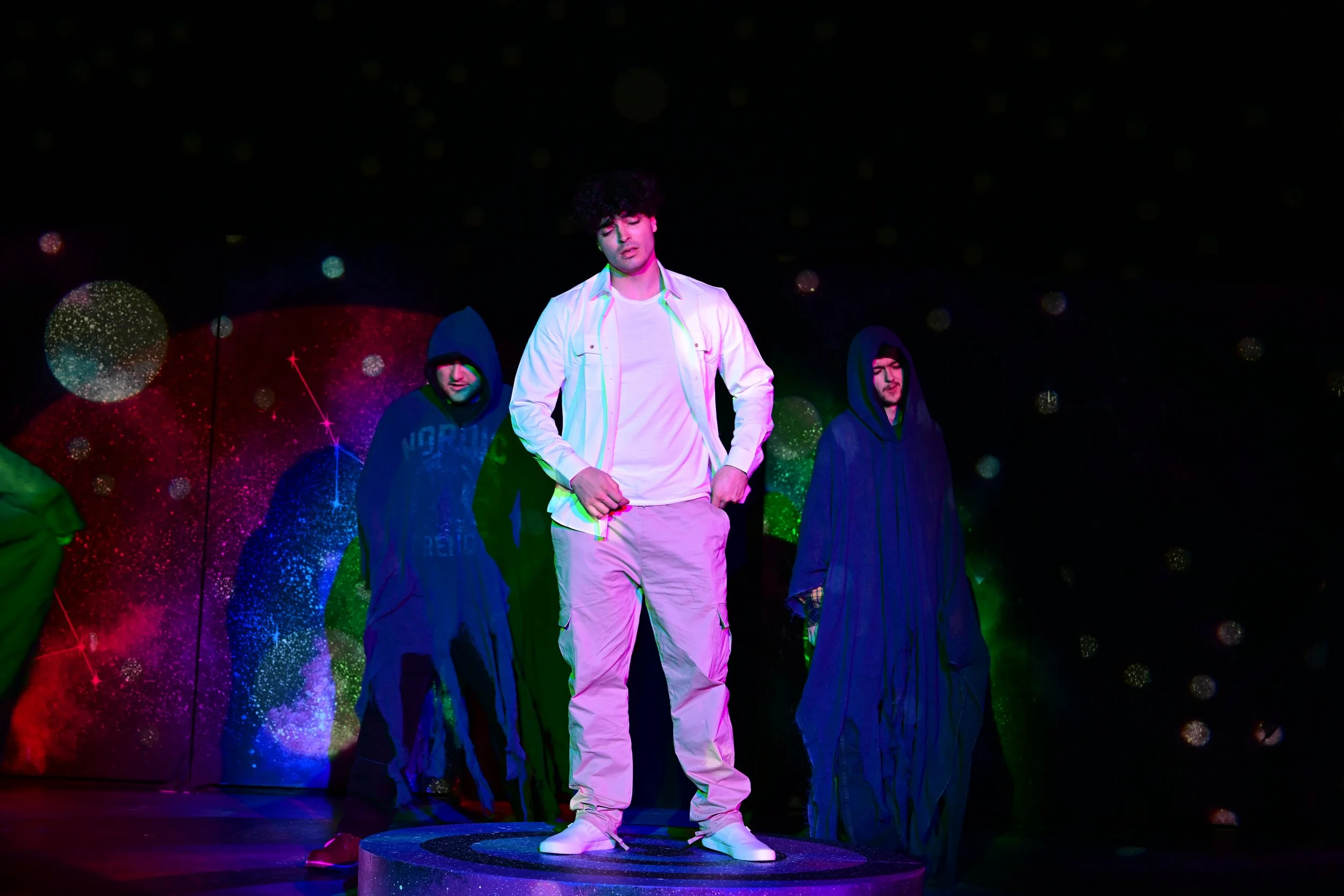 A young man with curly hair standing on a small platform with other people in dark hooded clothing behind him. The background features a multicolored cosmic or galaxy-themed display.