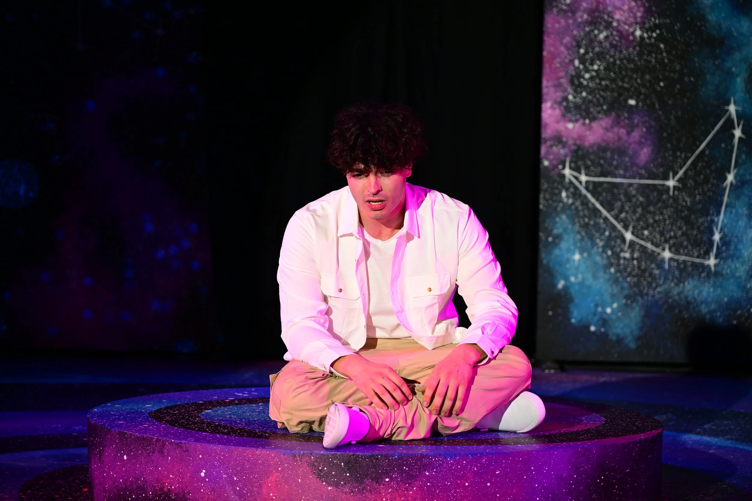 A person sitting cross-legged on a cosmic-themed stage with a backdrop of stars and constellations, wearing a white jacket and beige pants, with an emotional expression.