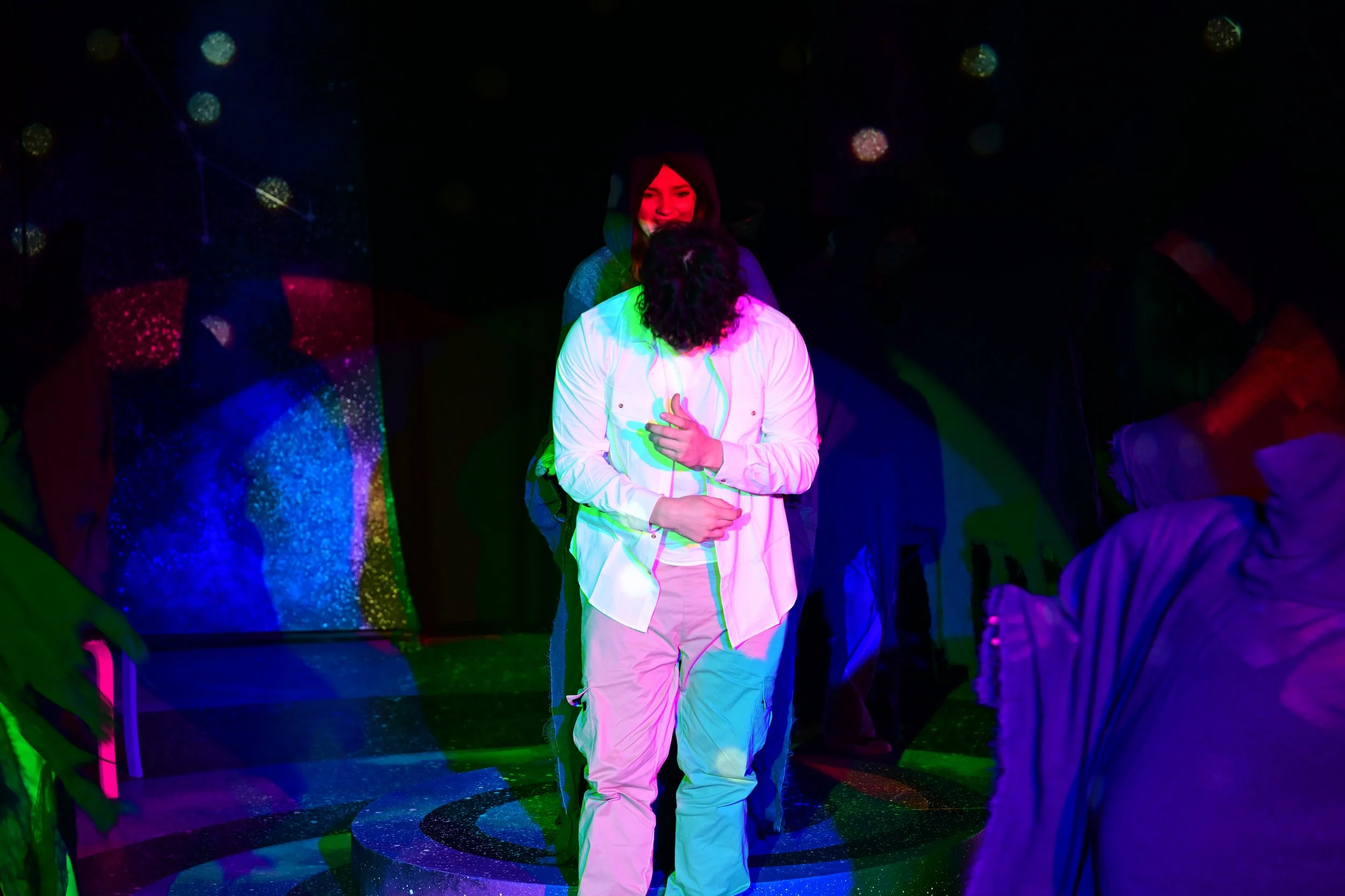 People in a dark room with colorful, glowing lights and star patterns, with one person standing and embracing another person's head.