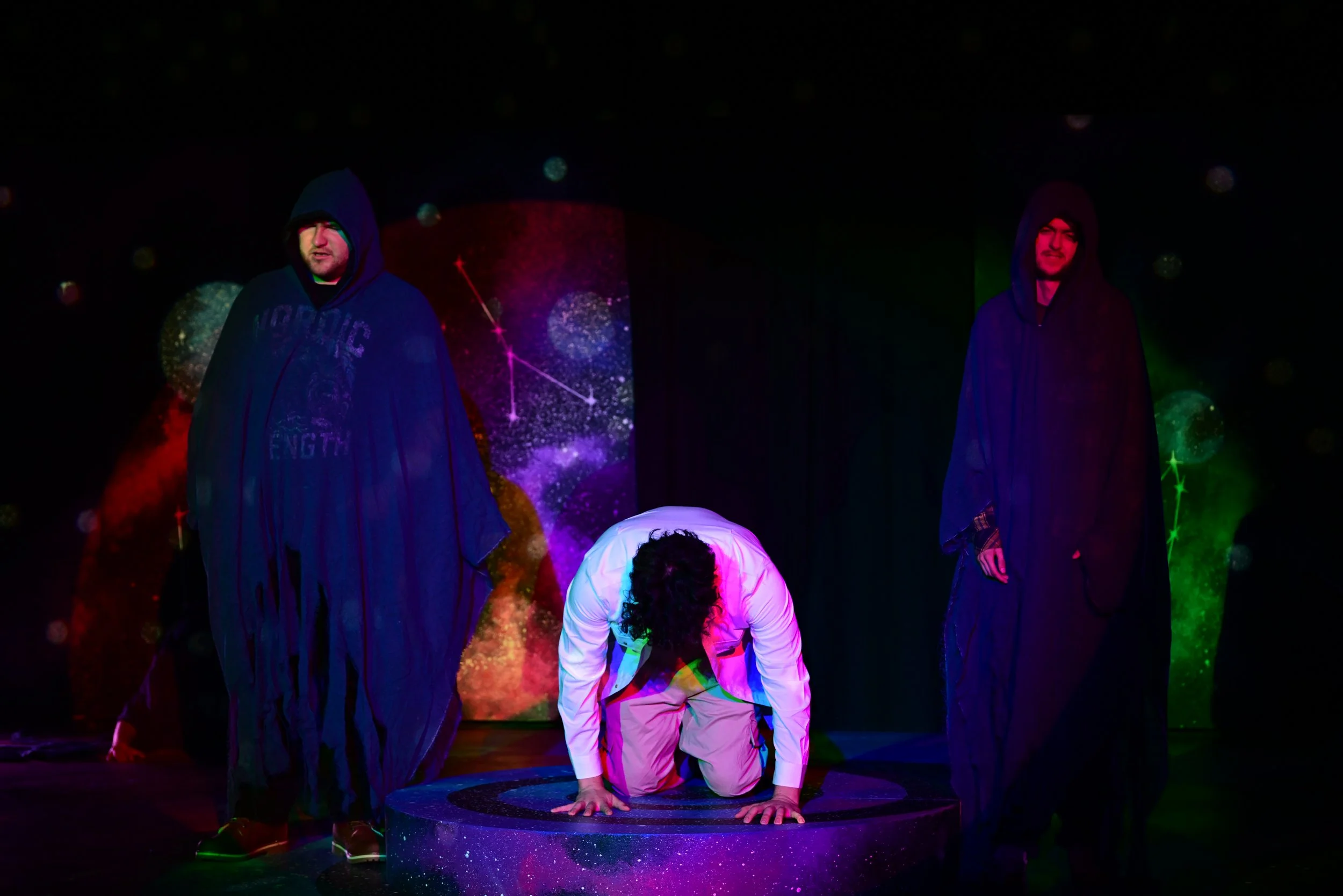 Stage performance with three actors; one kneeling and bowing on a round, starry platform, two standing wearing dark hooded cloaks; colorful cosmic-themed backdrop with stars and nebulae.