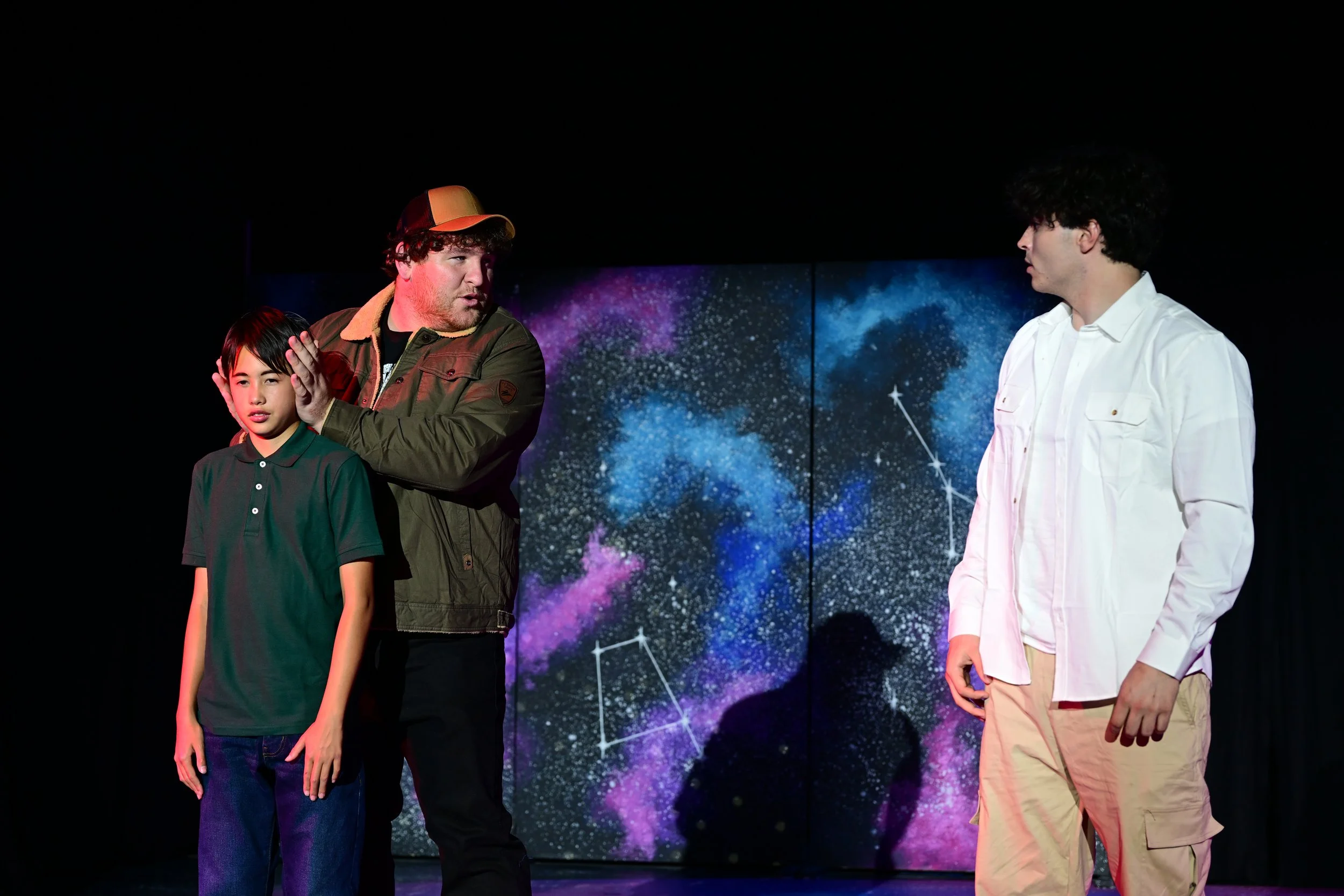 Three people standing on stage with a starry galaxy backdrop; one young boy with black hair in a green polo, a man in a cap and jacket gesturing, and another young man in a white shirt and beige pants listening.