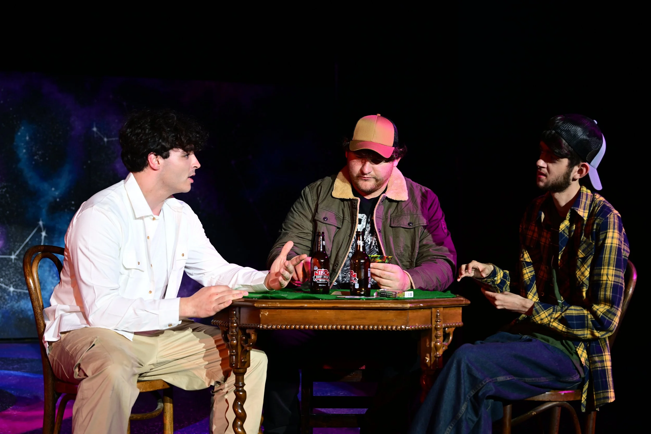 Three men sitting at a wooden table having a conversation with two beer bottles on the table. The man on the left is wearing a white shirt, the man in the middle is wearing a cap and a jacket, and the man on the right is wearing a plaid shirt and a b