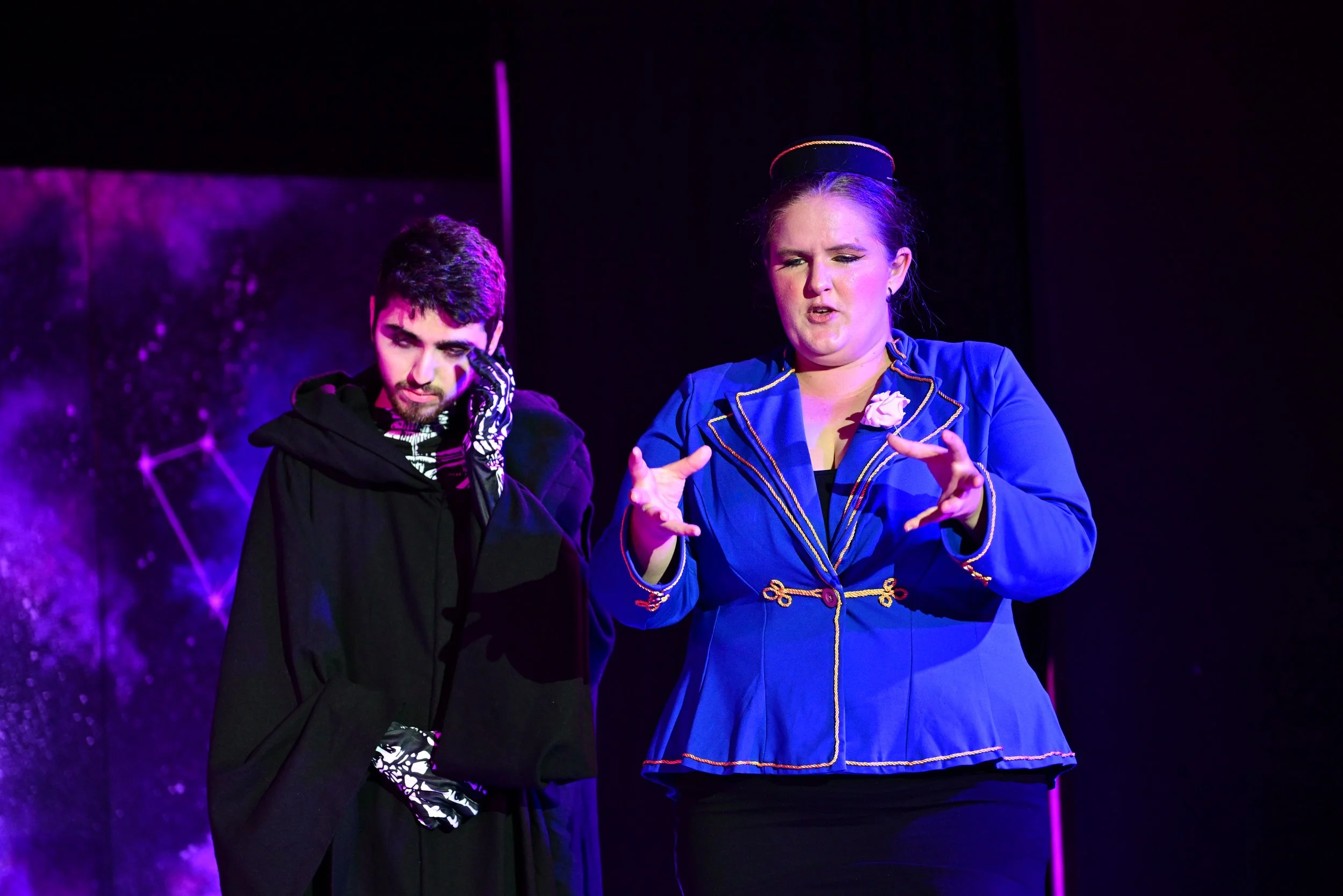 Two actors on stage, one man with dark hair and beard wearing a black cloak, and one woman with her hair in a bun wearing a royal blue costume with gold trim, performing a scene.