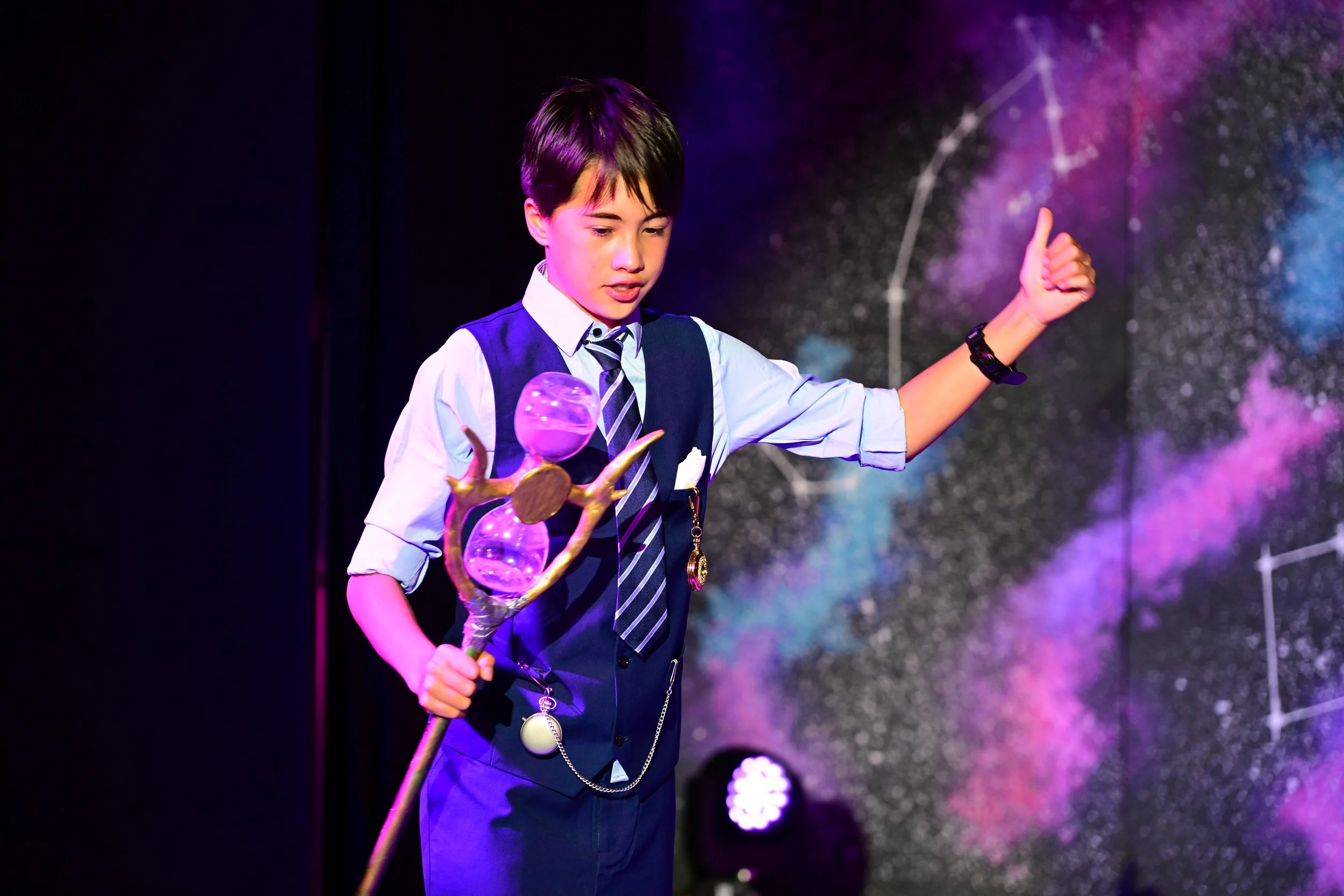 Young boy in school uniform performing on stage, holding a staff with a glowing purple orb, against a backdrop resembling a starry galaxy.
