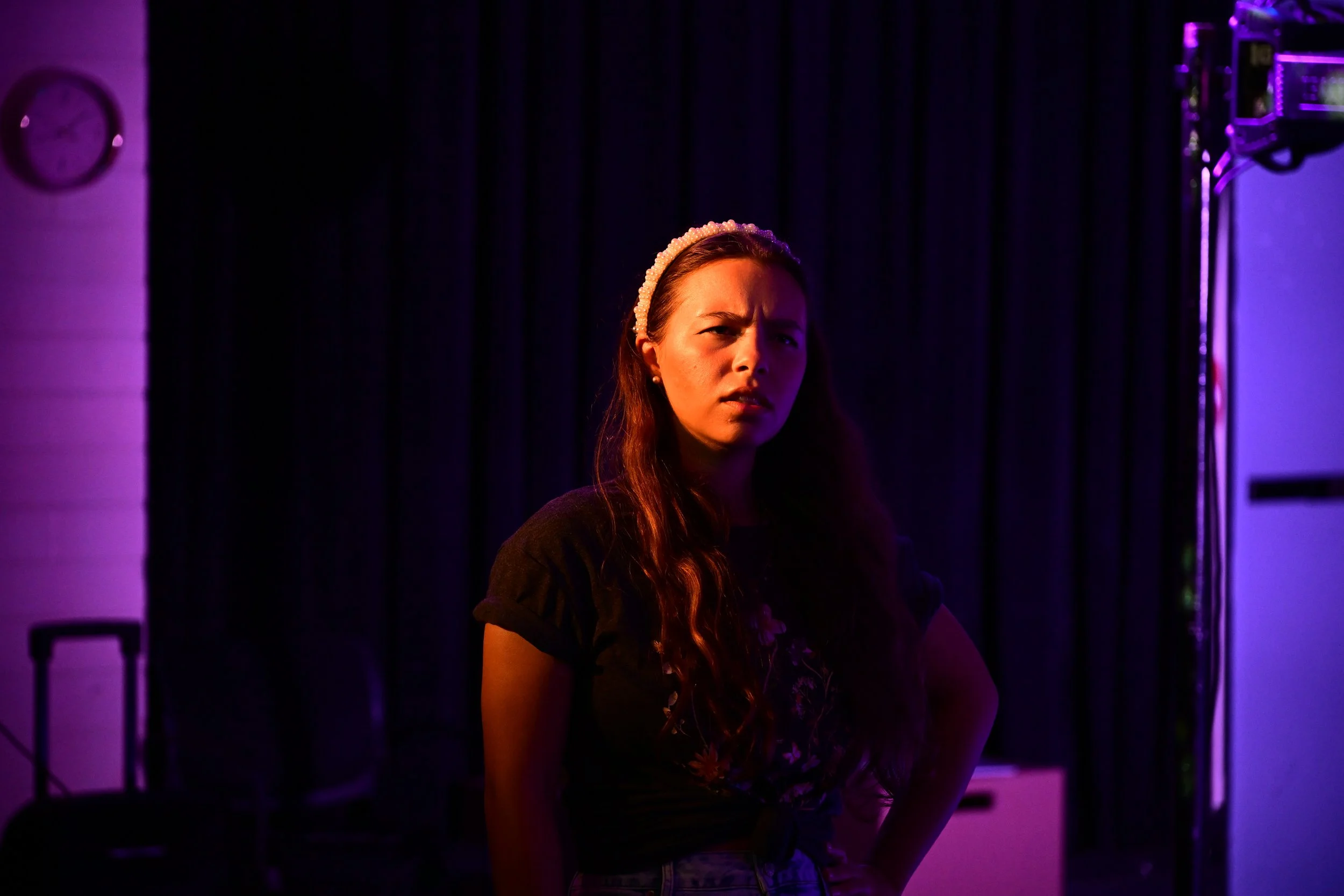 A woman stands with her hands on her hips in a dimly lit room with purple and blue lighting, dark curtain backdrop, and stage equipment visible.