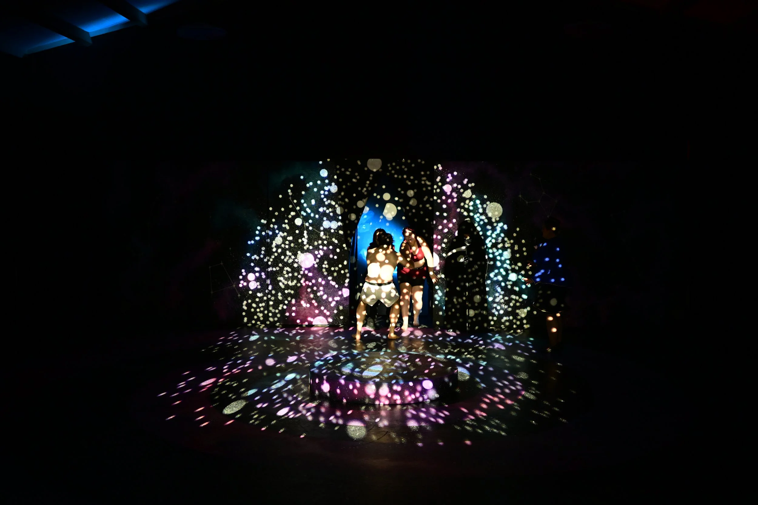 People standing on a stage with colorful, dotted light projections creating a starry effect around them at a dark event or party.