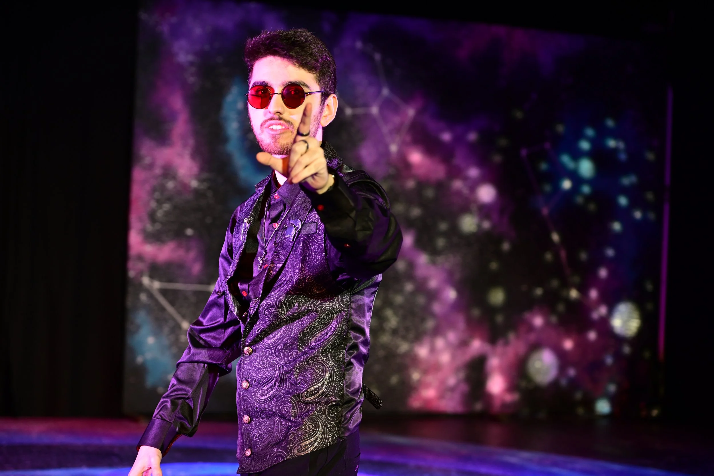 Person on stage wearing purple jacket with paisley pattern, red sunglasses, pointing towards camera, with a galaxy-themed backdrop.