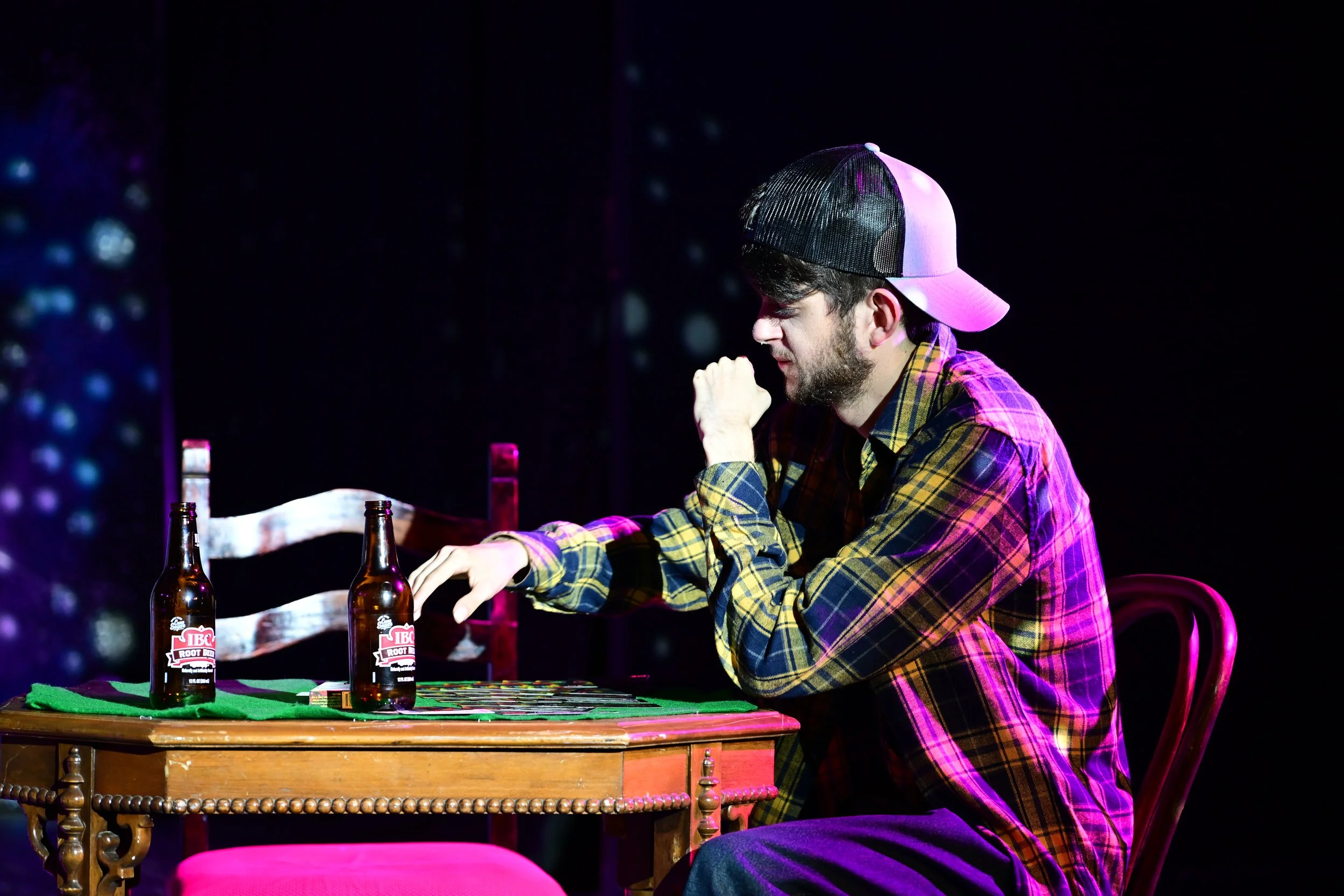 A man wearing a plaid shirt and baseball cap playing an intense game of chess at a table with two beer bottles during a performance or show.