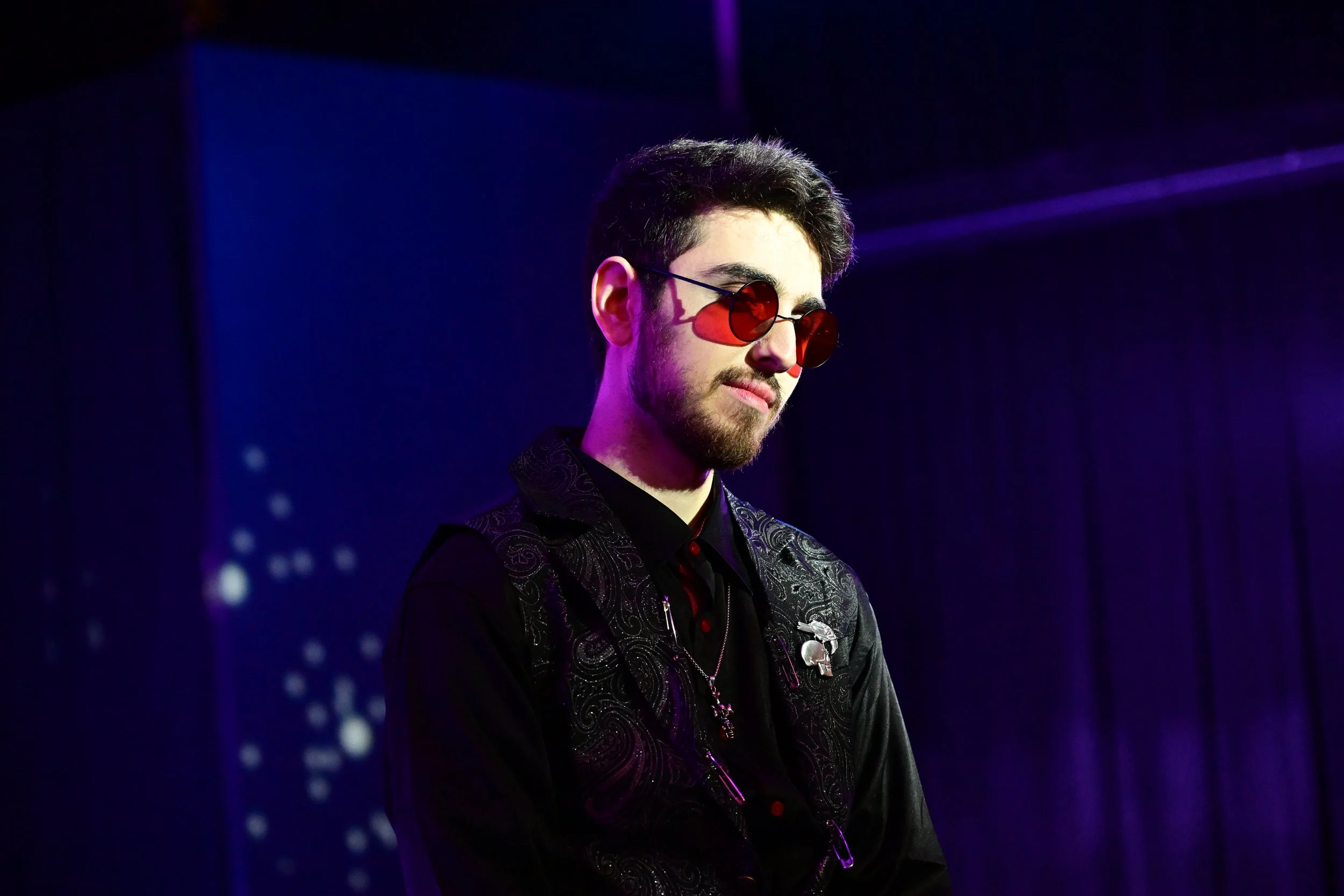 A young man with dark hair, wearing red sunglasses, a black shirt, and a black embroidered vest, stands in front of a dark background with purple lighting.