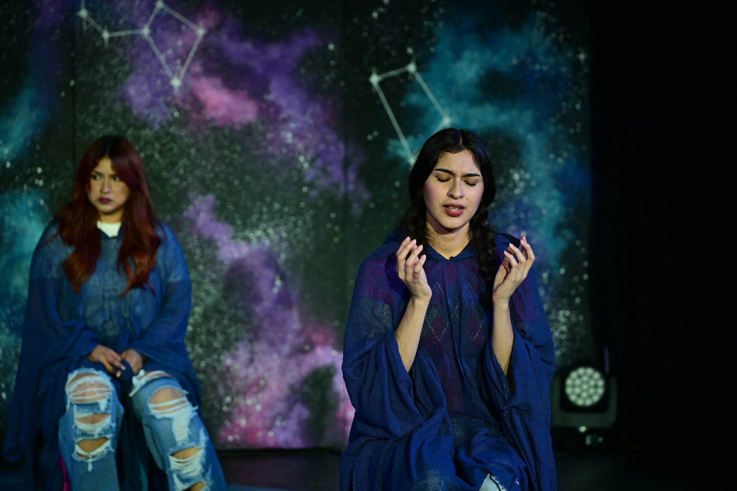 Two women in blue costumes with galaxy and star constellations backdrop, one with an emotional expression and the other looking pensive.