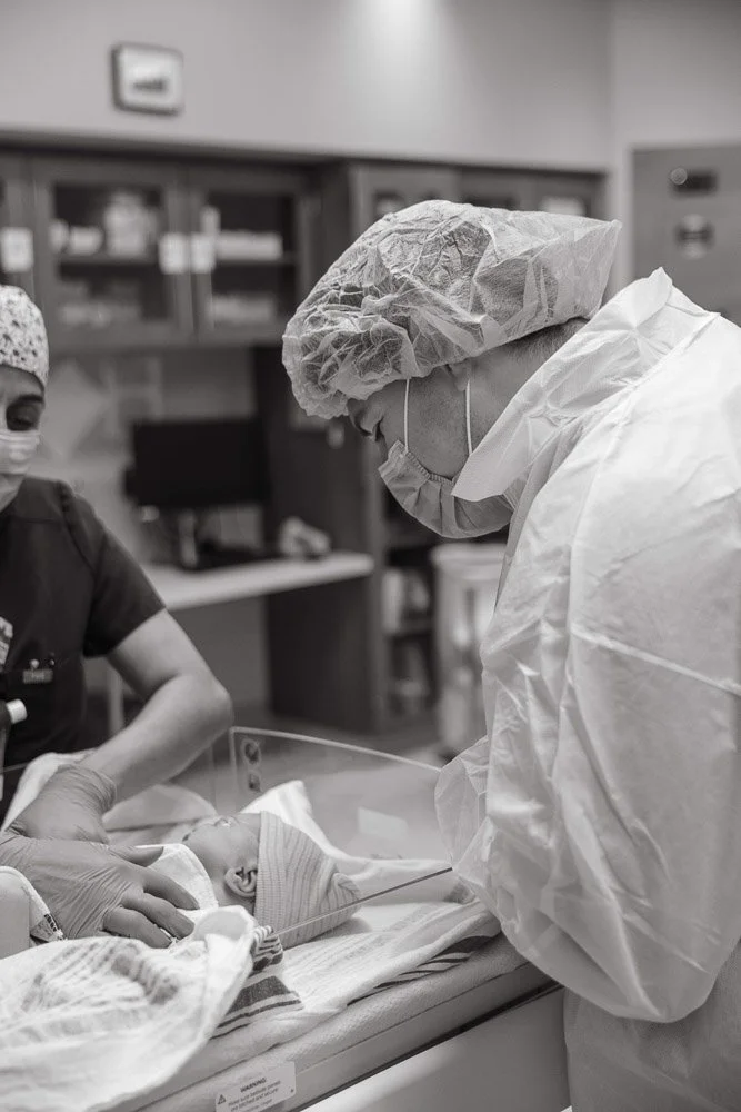 A healthcare worker in protective gear, including a hair cap, mask, and gown, tending to a newborn baby in a hospital setting.
