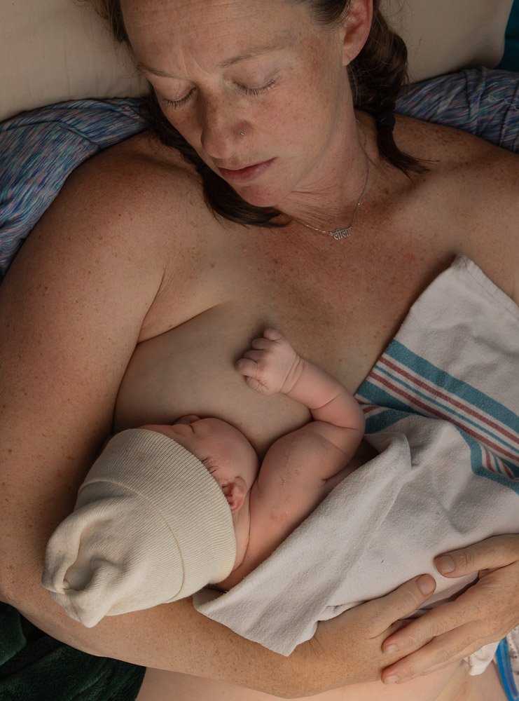 A woman breastfeeding an infant in a hospital bed, the woman is wearing a necklace and has a nose piercing, with her eyes closed.