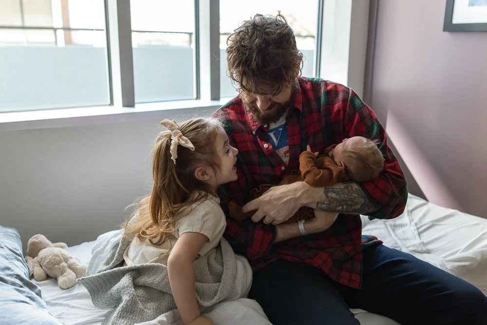 A man holding a newborn baby on a bed with a young girl, all inside a room with large windows.