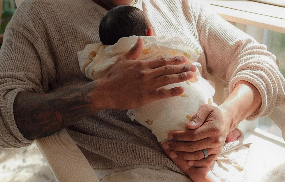 Person holding a newborn baby wrapped in a blanket, sitting by a window.