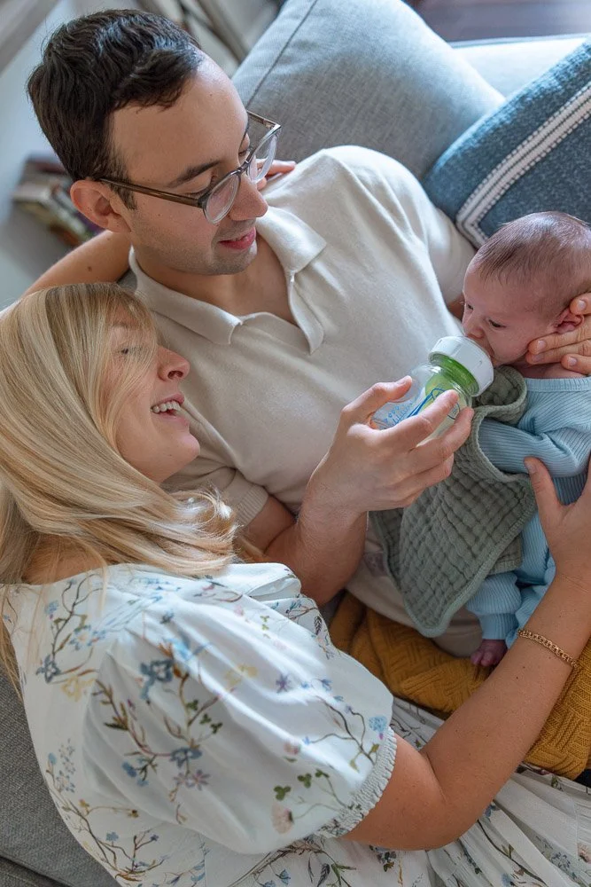 A family with a mother, father, and a baby lying on a couch, sharing a moment with the father feeding the baby with a bottle.